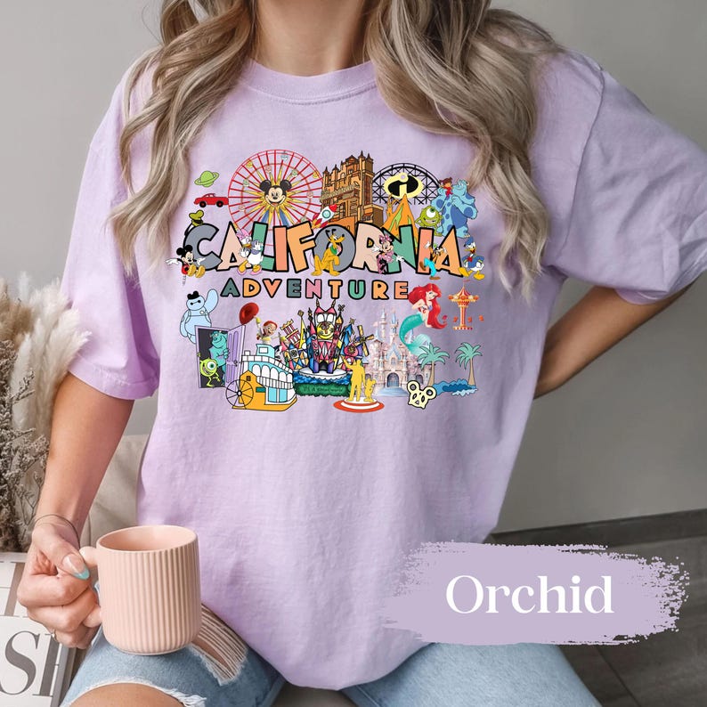 May include: A light purple t-shirt with a colorful graphic featuring the words "California Adventure" and various cartoon characters and landmarks. The shirt has short sleeves and a relaxed fit. The word "Orchid" is printed on the bottom right.