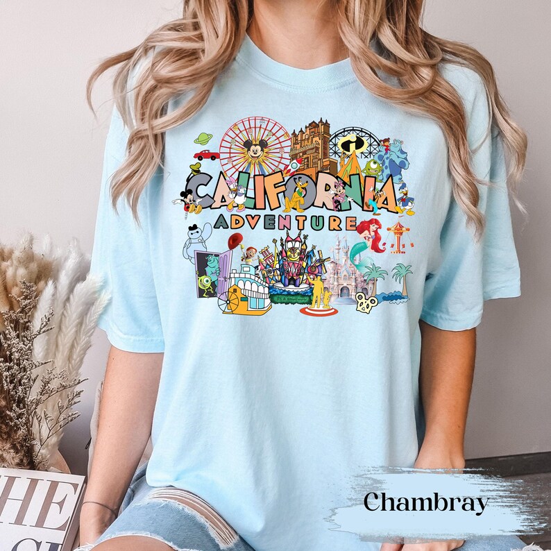 May include: Light blue t-shirt featuring a colorful graphic of the California Adventure theme park. The design includes a Ferris wheel, cartoon characters, and the words "CALIFORNIA ADVENTURE". The shirt is made of soft material.