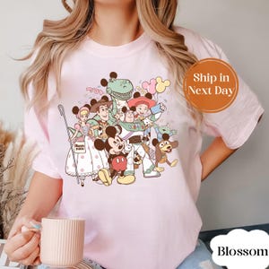 Może przedstawiać: Jasnoróżowa koszulka z kolorową grafiką postaci Disneya, w tym Myszki Miki, Chudego i Buzz'a Astrala. Projekt zawiera konfetti i balony. Koszulka ma okrągły dekolt i krótkie rękawy.