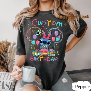 May include: Dark gray t-shirt featuring a cartoon character with a pink bow, surrounded by balloons and gifts. The shirt has the words "Custom Birthday" in colorful text. The design includes a birthday cake and stars.