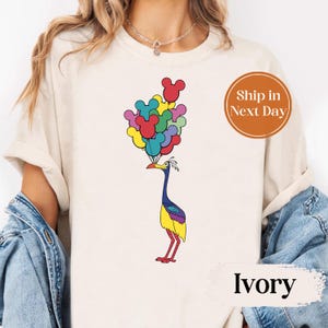 May include: Ivory-colored t-shirt featuring a colorful cartoon bird holding a bunch of Mickey Mouse-shaped balloons. The balloons are in various colors, including red, yellow, and blue. The shirt has a crew neck and short sleeves.