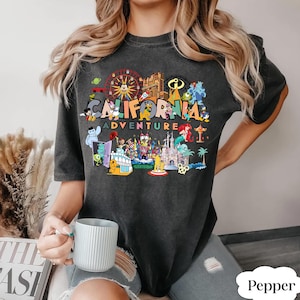 May include: Dark gray t-shirt with the words "CALIFORNIA ADVENTURE" in colorful letters. The shirt features a graphic of various theme park attractions and characters. The shirt is a relaxed fit.