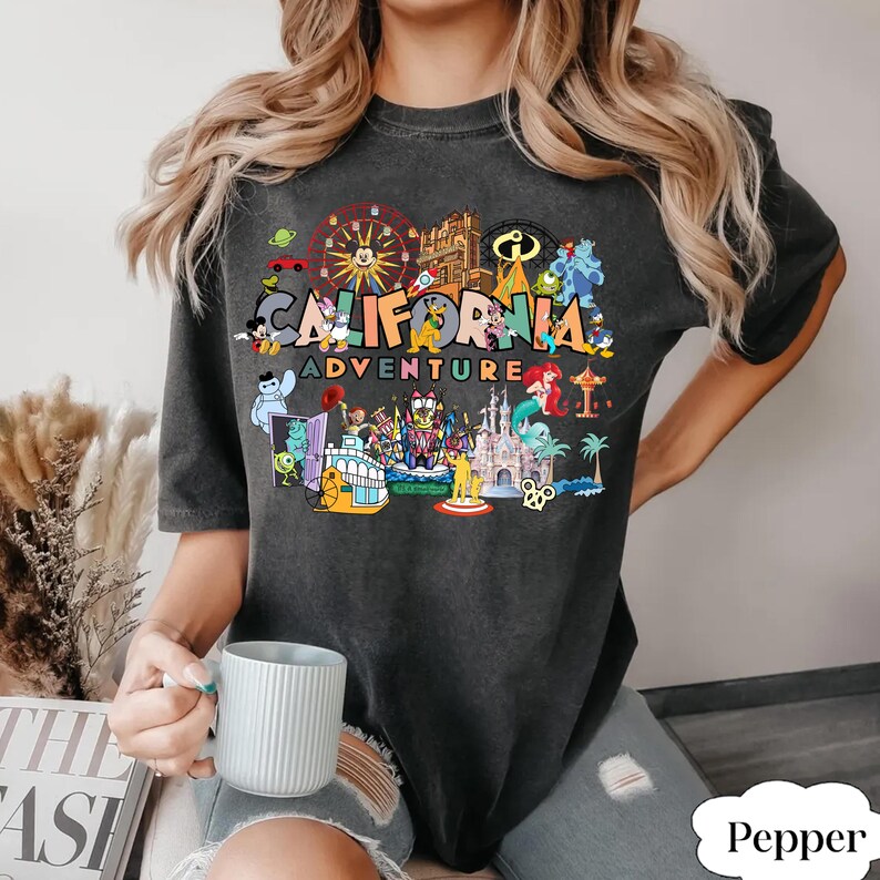May include: Dark gray t-shirt with the words "CALIFORNIA ADVENTURE" in colorful letters. The shirt features a graphic of various theme park attractions and characters. The shirt is a relaxed fit.
