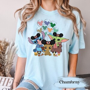 May include: Pale blue t-shirt with a vibrant graphic of Disney characters Stitch, Baby Groot, and Grogu, all wearing Mickey Mouse ears. The design includes balloons, popcorn, and food items. The word "Chambray" is printed at the bottom.