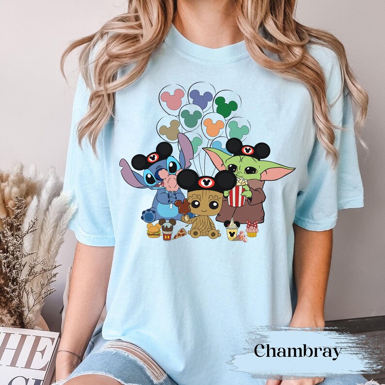 May include: Pale blue t-shirt with a vibrant graphic of Disney characters Stitch, Baby Groot, and Grogu, all wearing Mickey Mouse ears. The design includes balloons, popcorn, and food items. The word "Chambray" is printed at the bottom.