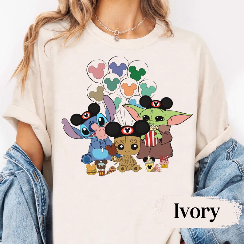 May include: Ivory-coloured t-shirt featuring a colourful graphic of Stitch, Baby Yoda, and Groot wearing Mickey Mouse ears and holding snacks. The characters are surrounded by Mickey Mouse balloon designs. The word "Ivory" is printed at the bottom.