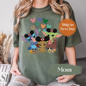 May include: Moss green t-shirt with a graphic of Stitch, Baby Groot, and Grogu wearing Mickey Mouse ears. The characters are holding food and balloons. The shirt has the word "Moss" printed on it.