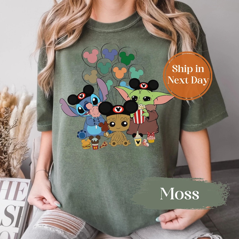 May include: Moss green t-shirt with a graphic of Stitch, Baby Groot, and Grogu wearing Mickey Mouse ears. The characters are holding food and balloons. The shirt has the word "Moss" printed on it.