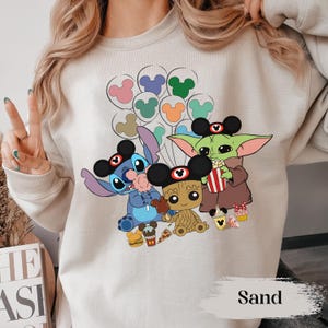 May include: Sand-coloured sweatshirt featuring cartoon characters Stitch, Baby Yoda, and Groot wearing Mickey Mouse ears. The design includes Mickey Mouse balloon graphics and food items like popcorn and cupcakes. The word "Sand" is printed at the bottom.