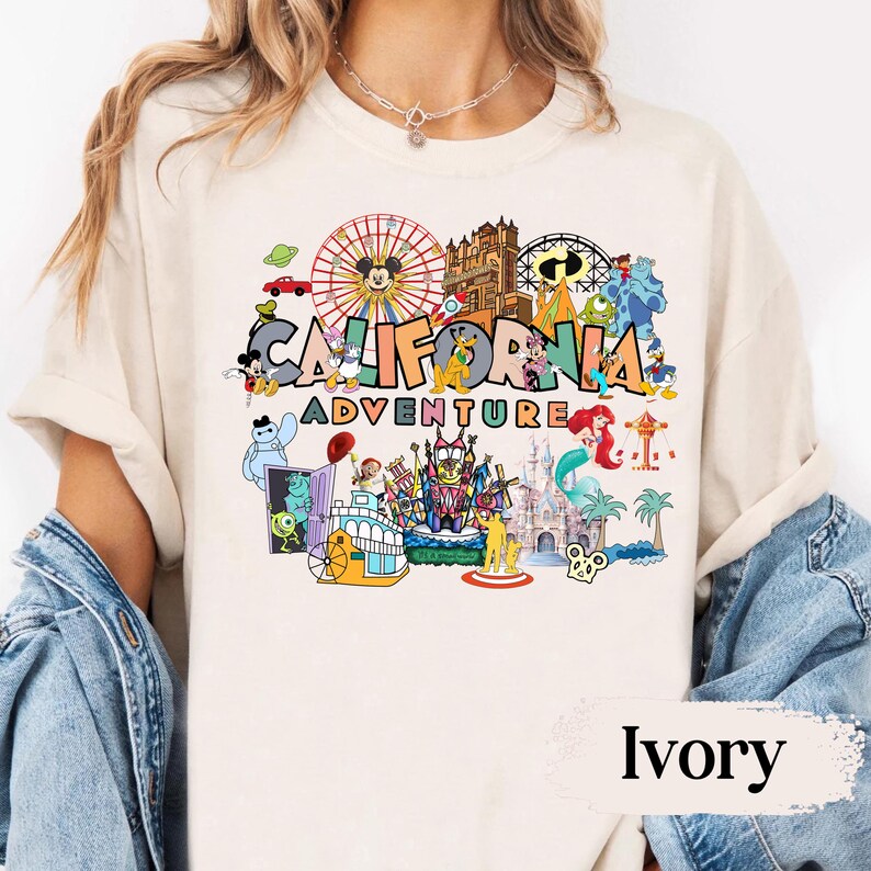 May include: Ivory-colored t-shirt featuring a colorful graphic of the California Adventure theme park. The design includes a Ferris wheel, cartoon characters, and the words "CALIFORNIA ADVENTURE". The shirt is made of a soft material.