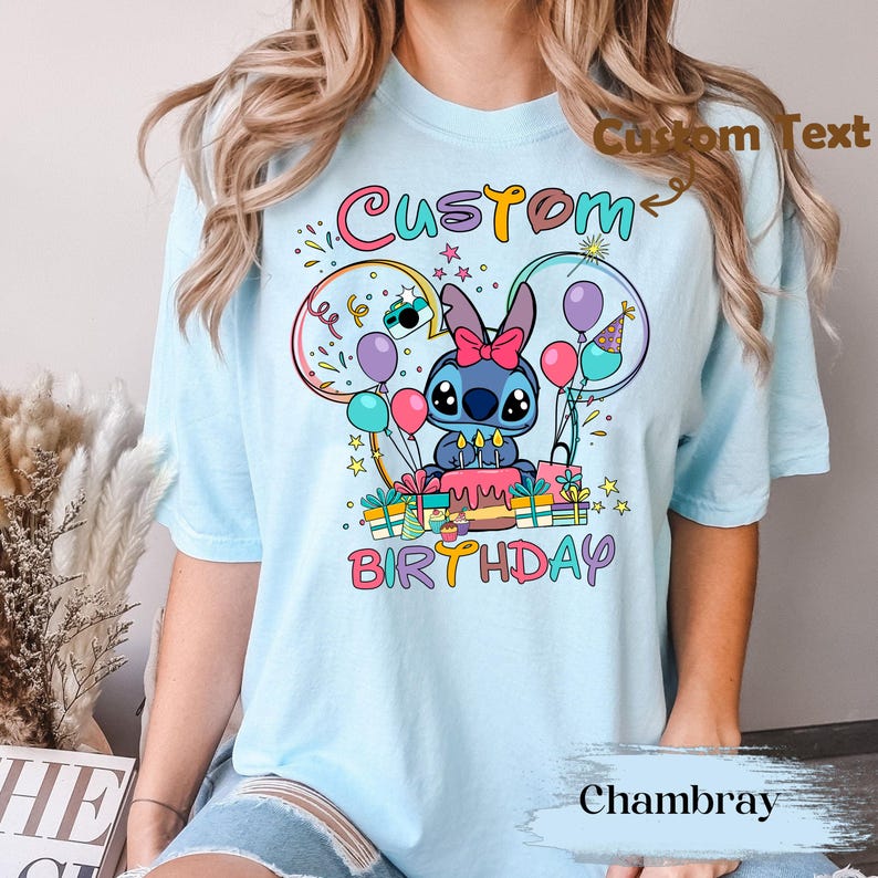 May include: Light blue t-shirt featuring a cartoon character with a pink bow, surrounded by balloons and gifts. The shirt has the words "Custom Birthday" in colorful text. The shirt is made of chambray fabric.