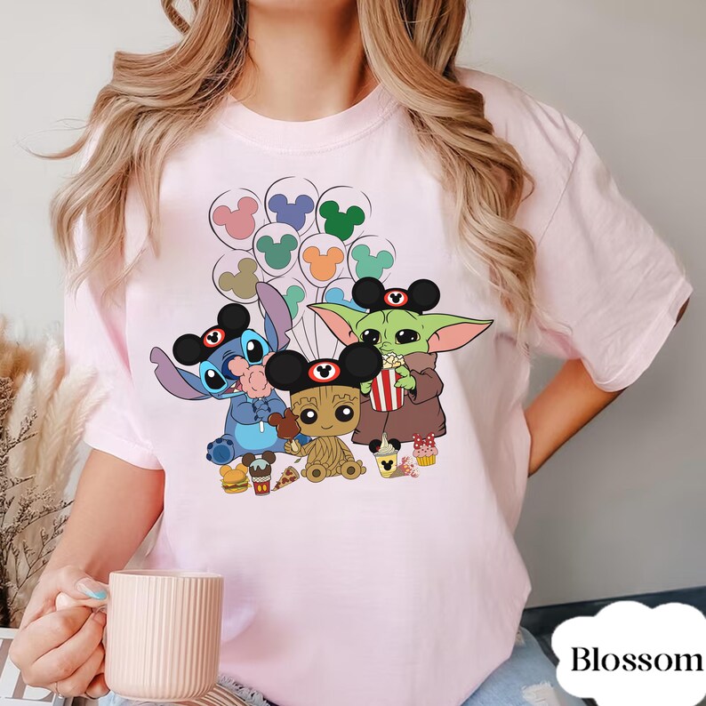 May include: A pale pink t-shirt with a cartoon graphic of Stitch, Baby Groot, and Grogu, all wearing Mickey Mouse ears. The characters are holding snacks and balloons. The word "Blossom" is printed on the bottom right.