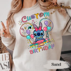 May include: Sand-colored sweatshirt featuring a colorful graphic of a blue cartoon character with a pink bow, surrounded by balloons, confetti, and the words "Custom Birthday." The design includes a birthday cake and wrapped gifts.