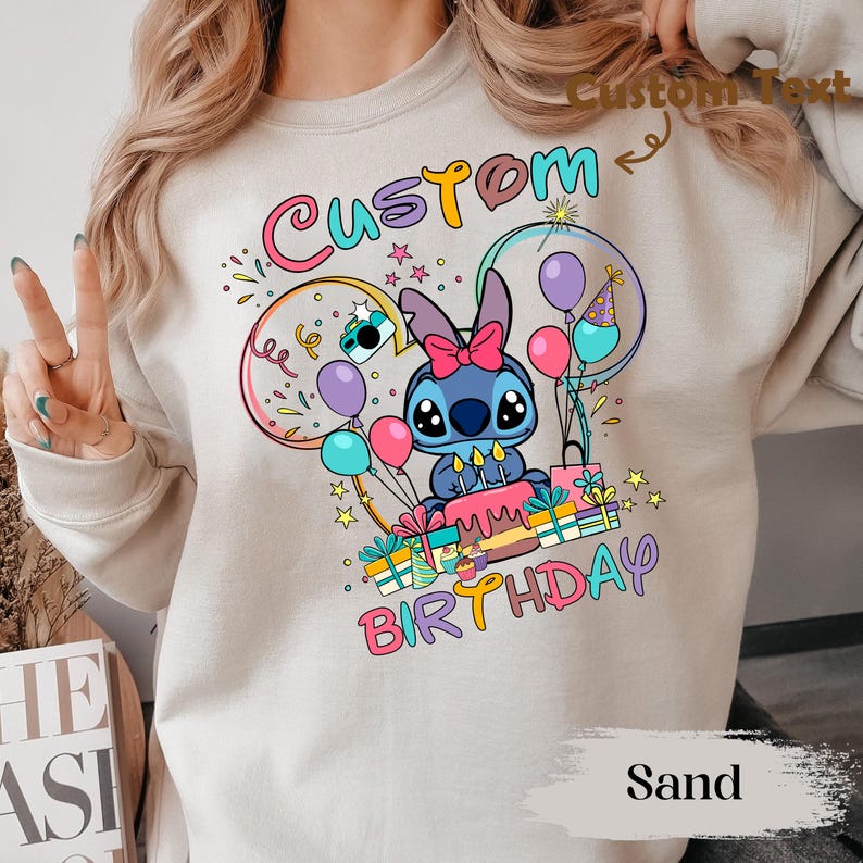 May include: Sand-colored sweatshirt featuring a colorful graphic of a blue cartoon character with a pink bow, surrounded by balloons, confetti, and the words "Custom Birthday." The design includes a birthday cake and wrapped gifts.