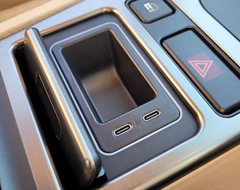Puede incluir: Primer plano del interior de un coche que muestra un compartimento plateado y negro con una tapa deslizante. El compartimento tiene dos puertos USB y una pequeña abertura rectangular. El compartimento está situado junto a un botón con un símbolo de triángulo rojo.
