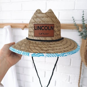 May include: A straw hat with a wide brim, featuring a brown leather patch with the text "LINCOLN crew." The brim is trimmed with a blue and white wave pattern. The hat has a black chin strap.