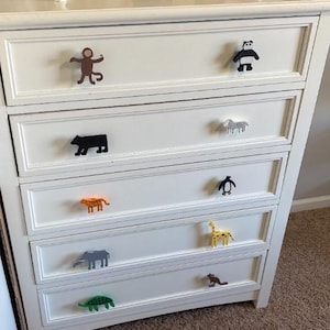 May include: A white dresser with animal-shaped drawer pulls. The pulls include a monkey, panda, bear, zebra, tiger, penguin, giraffe, elephant, kangaroo, and turtle. The dresser has five drawers.