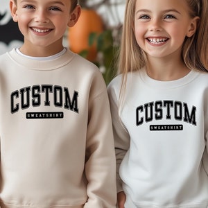 May include: Two children wearing matching sweatshirts with the text "CUSTOM SWEATSHIRT" in black. The boy is wearing a beige sweatshirt and the girl is wearing a white sweatshirt. They are standing in front of a white brick wall with pumpkins.