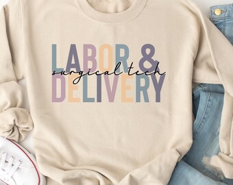 Op de afbeelding: Een beige sweatshirt met de tekst "LABOR & surgical tech DELIVERY" in kleurrijke letters. Het sweatshirt ligt op een wit oppervlak met een paar witte sneakers en een blauwe jeans.