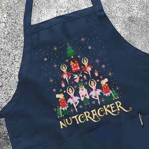 May include: Navy blue kitchen apron with a Christmas tree design featuring a Nutcracker theme. The tree is decorated with dancers, a nutcracker, and other festive elements. The text "NUTCRACKER" is printed in gold at the bottom of the design.