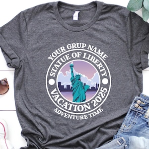 May include: Dark gray t-shirt with a circular graphic featuring the Statue of Liberty, text that reads "Your Group Name, Statue of Liberty, Vacation 2025, Adventure Time." Includes blue jeans, white sneakers, and sunglasses.