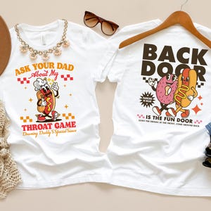 Puede incluir: Dos camisetas blancas con gr&aacute;ficos humor&iacute;sticos. Una dice "Ask Your Dad About My Throat Game" con un perrito caliente de dibujos animados. La otra dice "Back Door Is The Fun Door" con un cerebro y un perrito caliente de dibujos animados.