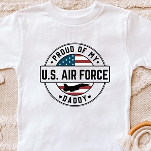 May include: White t-shirt featuring a circular design with the text "PROUD OF MY U.S. AIR FORCE DADDY". The design includes an American flag and a silhouette of a jet. A patriotic themed tee.