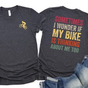 May include: Dark gray t-shirts with a bicycle graphic and the text "Sometimes I wonder if my bike is thinking about me too." White sneakers and blue jeans are also in the image.