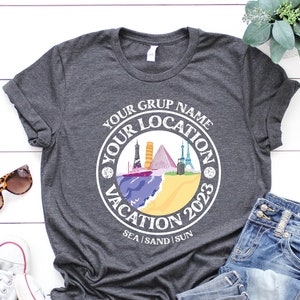 May include: A gray t-shirt with a white circle design featuring the text "Your Group Name Your Location Vacation 2023 Sea Sand Sun". The design also includes a colorful illustration of various landmarks.