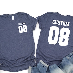 May include: Two navy blue t-shirts with the text "CUSTOM 08" in white, one on the front and one on the back. A pair of blue jeans and white trainers are also in the image. The shirts are likely for a sports team or a personalised gift.