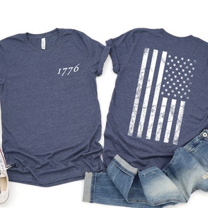 K&ouml;nnte beinhalten: Zwei marineblaue T-Shirts, eines mit der Zahl "1776" auf der Vorderseite und das andere mit einer wei&szlig;en, abgenutzten amerikanischen Flagge auf der R&uuml;ckseite.