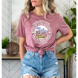 K&ouml;nnte beinhalten: Ein mauvefarbenes T-Shirt mit einem wei&szlig;en Kreis-Motiv, das eine Stadtlandschaft und den Text "Your Grup Name Your Location Vacation 2024 Adventure Time" zeigt.