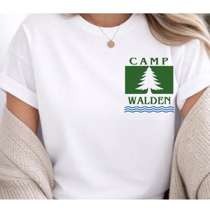 May include: White t-shirt with a green and white graphic. The graphic features the text "CAMP WALDEN" above a white tree and blue wavy lines. The shirt has rolled-up sleeves.
