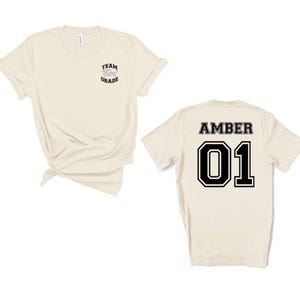 May include: Two cream-colored t-shirts. One shirt has "TEAM Third GRADE" printed on the front. The other shirt has "AMBER" and the number "01" printed on the back in black block letters with a white border. Both shirts are short-sleeved.