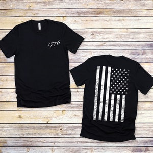 K&ouml;nnte beinhalten: Schwarzes T-Shirt mit der Zahl 1776 auf der Vorderseite und einem verwitterten amerikanischen Flaggenmuster auf der R&uuml;ckseite.