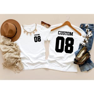 May include: Two white t-shirts, each with "CUSTOM" and the number "08" printed in black. One is on a hanger, the other flat. Accessories include a brown hat, sunglasses, and a necklace. Jeans and leopard print shoes are also visible.