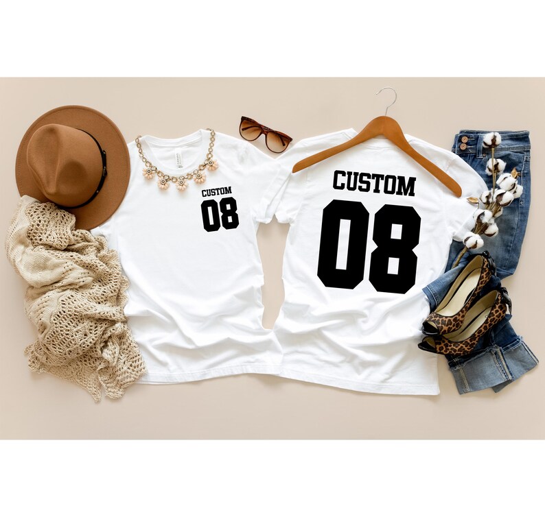 May include: Two white t-shirts, each with "CUSTOM" and the number "08" printed in black. One is on a hanger, the other flat. Accessories include a brown hat, sunglasses, and a necklace. Jeans and leopard print shoes are also visible.