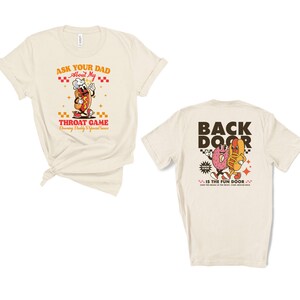 Puede incluir: Dos camisetas color crema. Una presenta un perrito caliente de dibujos animados con el texto "ASK YOUR DAD About My THROAT GAME Drawing Daddy's Special Sauce". La otra camiseta tiene un perrito caliente de dibujos animados y un donut de gelatina con el texto "BACK DOOR IS THE FUN DOOR."