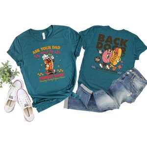 Puede incluir: Camisetas de color turquesa con gr&aacute;ficos de dibujos animados de perritos calientes y donuts. Una camiseta dice "Ask Your Dad About My Throat Game" y la otra "Back Door". Las camisetas se combinan con vaqueros azules y zapatillas blancas.