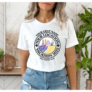 May include: White t-shirt with a black circle graphic that says "Your Group Name Your Location Vacation 2023 Sea | Sand | Sun". The graphic features a colorful illustration of a beach scene with buildings and a boat.