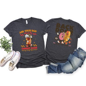 Puede incluir: Camisetas de color gris oscuro con gr&aacute;ficos delanteros y traseros. La parte delantera presenta un perrito caliente de dibujos animados con el texto "Ask Your Dad About My Throat Game". La parte trasera muestra un donut y un perrito caliente con el texto "Back Door."