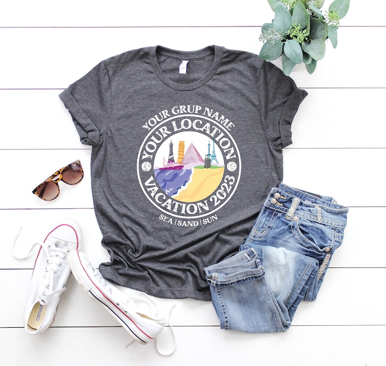 May include: A gray t-shirt with a white circle design featuring the text "Your Group Name Your Location Vacation 2023 Sea Sand Sun". The design also includes a colorful illustration of various landmarks.