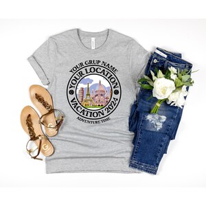K&ouml;nnte beinhalten: Graues T-Shirt mit einem schwarzen Kreis-Motiv, auf dem "Your Grup Name Your Location Vacation 2024 Adventure Time" steht. Das Motiv zeigt eine Stadtlandschaft mit Geb&auml;uden und blauem Himmel.