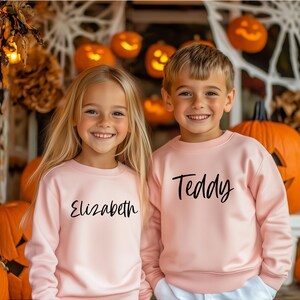 Peut inclure: Deux enfants portant des sweat-shirts roses avec leurs noms, "Elizabeth" et "Teddy", imprim&eacute;s en noir. Ils sont debout devant un fond de citrouilles et de d&eacute;corations d'automne.