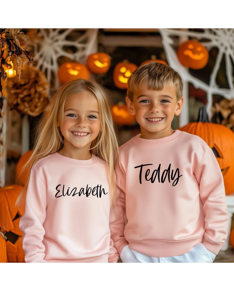 Peut inclure: Deux enfants portant des sweat-shirts roses avec leurs noms, "Elizabeth" et "Teddy", imprim&eacute;s en noir. Ils sont debout devant un fond de citrouilles et de d&eacute;corations d'automne.