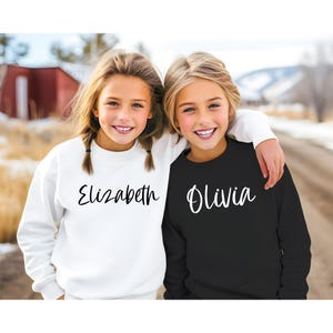 Peut inclure: Deux filles portant des sweat-shirts assortis avec leurs noms, Elizabeth et Olivia, &eacute;crits en &eacute;criture noire sur le devant. La fille &agrave; gauche porte un sweat-shirt blanc et la fille &agrave; droite porte un sweat-shirt noir.