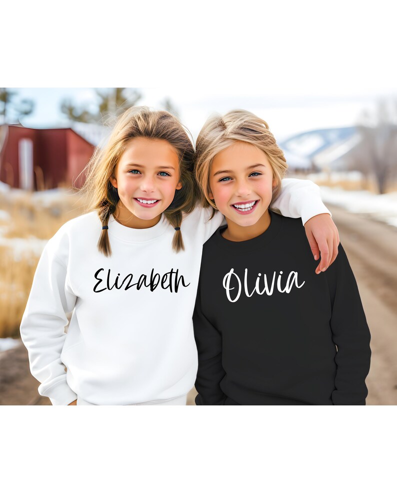 Peut inclure: Deux filles portant des sweat-shirts assortis avec leurs noms, Elizabeth et Olivia, &eacute;crits en &eacute;criture noire sur le devant. La fille &agrave; gauche porte un sweat-shirt blanc et la fille &agrave; droite porte un sweat-shirt noir.