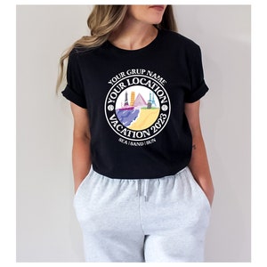 May include: Black t-shirt with a white circle graphic featuring the text "Your Grup Name Your Location Vacation 2023 Sea | Sand | Sun" and a colorful illustration of various landmarks.