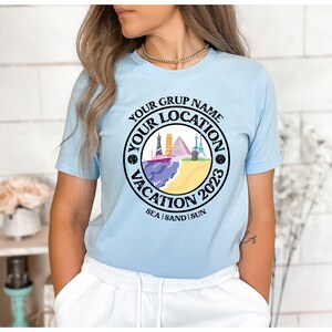 May include: Light blue t-shirt with a black circle graphic featuring a beach scene with buildings, a ship, and the text "Your Grup Name Your Location Vacation 2023 Sea | Sand | Sun".