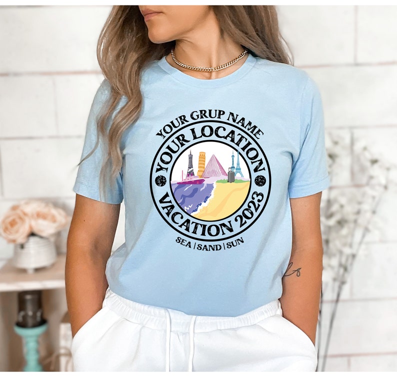 May include: Light blue t-shirt with a black circle graphic featuring a beach scene with buildings, a ship, and the text "Your Grup Name Your Location Vacation 2023 Sea | Sand | Sun".